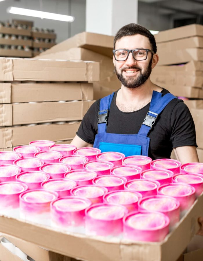 Uśmiechnięty pracownik w magazynie, ubrany w niebieskie ogrodniczki, trzymający tacę z różowymi opakowaniami produktów, otoczony stosami kartonowych pudeł.